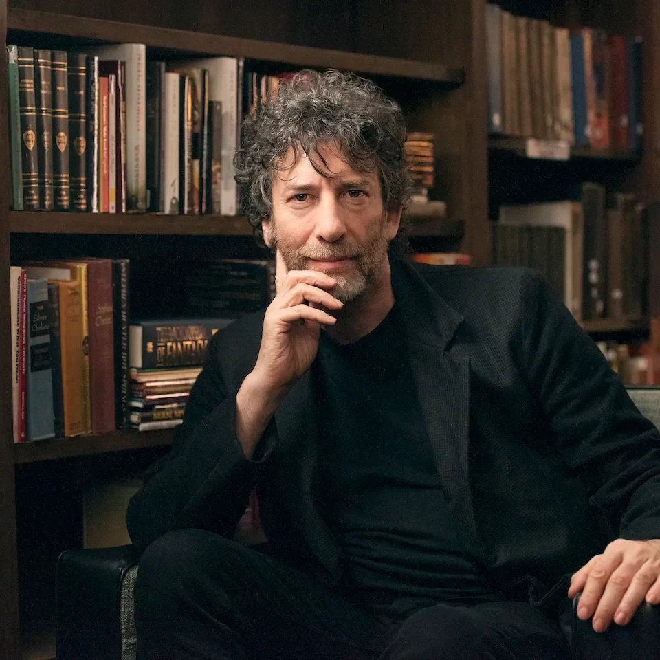 Neil Gaiman sat in a library wearing a dark suit ad top
